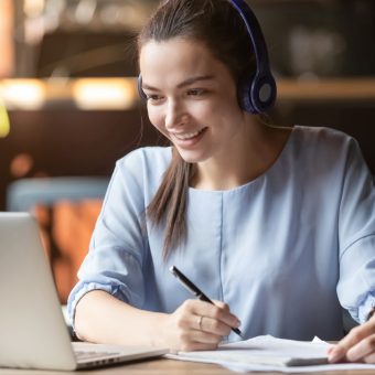 Smiling girl student wear wireless headphone study online with skype teacher, happy young woman learn language listen lecture watch webinar write notes look at laptop sit in cafe, distant education
